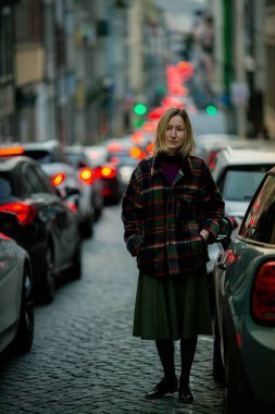 Akşamları yoğun, trafik dolu bir şehir sokağında bir kadın duruyor. Etrafı far ve kentsel hayatla çevrili..