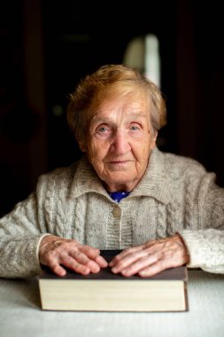 Yaşlı bir kadın, evinde bir masada otururken, elleri kalın bir kitabın üzerinde dinlenen bir portre için poz veriyor..
