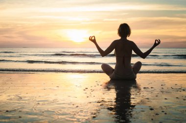 Yoga yapan bir kadının silueti gün batımında plajda poz vererek huzurlu ve meditasyon yapan bir atmosfer yaratıyor..