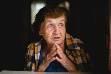 Evde bir masada oturan yaşlı bir kadının portresi. Elleri sımsıkı sarılmış ve düşünceli ifadesine yansıyor..