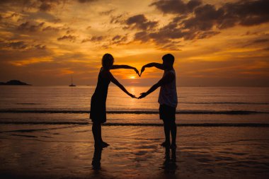 Alacakaranlıkta deniz kıyısındaki bir kumsaldaki bir çiftin silueti, elleriyle batan güneşin arka planına karşı bir kalp oluşturuyor..