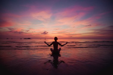 Sahilde gerçek üstü bir günbatımına karşı nilüfer çiçeğiyle yoga yapan bir kadının silüeti..