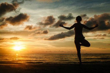 Sihirli bir günbatımı sırasında tropik bir plajda yoga yapan bir kadının silueti..