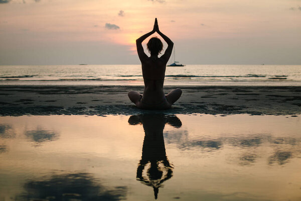 Силуэт женщины в позе лотоса, практикующей йогу на тропическом пляже в сумерках в Таиланде, ее отражение мерцает в спокойной воде.
