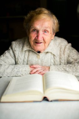 Yaşlı bir kadın bir masada oturur ve kalın bir kitap okur. Etrafı bilgelik ve sakinlik duygusuyla çevrili..