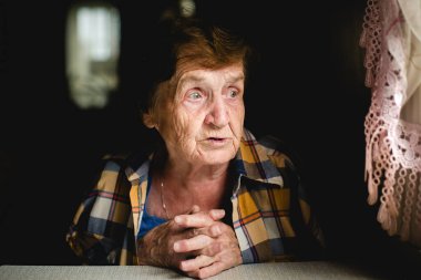 Yaşlı bir kadın sessizce oturur, elleri bağlı, düşünceli bir şekilde pencereye bakar. Yüzü sakin ve odaklanmış.