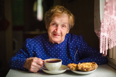 Yaşlı bir kadın evinde huzur içinde çay içiyor, yüzü sakin ve memnun. Portre, konfor, sadelik ve sessiz sevinçle dolu nazik, sıcak bir anı yakalıyor..