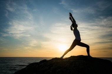 Gün batımında deniz kenarında jimnastik yapan bir kadının silueti. Zarif hareketleri, parlayan gökyüzü ve dalgaların sakin ritmi tarafından çerçevelenir..