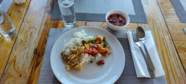 Pirinç, karışık sebze ve kızarmış yiyeceklerin tepeden aşağı manzarası, yanında bir kase tatlı çorbası, bir bardak su ve ahşap masa seti üzerindeki çatal bıçak takımı; mutfak, akşam yemeği veya kültürel gıda deneyimleri kavramları.