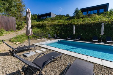 Açık havada, açık mavi bir gökyüzünün altında şezlonglu sandalyeli ve şemsiyeli bir yüzme havuzu. Havuz alanı çimenli bir tepede, arka plandaki yamaçta modern kamaralar görülebilir..