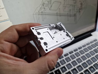 Mechanical engineer hand holding CNC milled custom designed project in front of computer screen and drawings