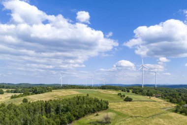 Parlak mavi gökyüzüne karşı yemyeşil tarlalar ve rüzgar türbinlerinin yer aldığı pitoresk bir manzara, yenilenebilir enerji ve doğal güzellik sergiliyor..