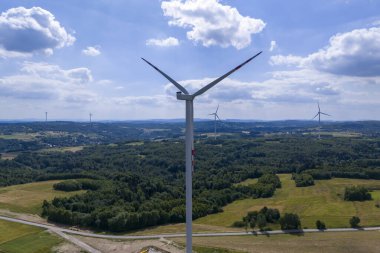 Pofuduk bulutlu açık mavi gökyüzünün altında, verimli yeşil bir arazide rüzgar türbinlerinin hava görüntüsü sürdürülebilir enerji üretimi sergiliyor..