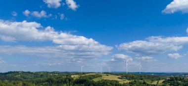 Tüylü bulutlu mavi bir gökyüzünün altında yeşil bir kırsalda rüzgar türbinleri barındıran güzel bir manzara. Çevre dostu temalar ve yenilenebilir enerji kavramları için mükemmel.