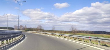 Mavi gökyüzünün altında, ağaçlar ve tarlalarla çevrili, güneşli bir günde doğanın güzelliğini yakalayan, dolambaçlı bir yolun manzarası.