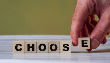 Bir el 'E' harfini tahta blokların yanına 'CHOOS' yazarak görsel olarak seçenekleri ve kararları temsil ediyor..