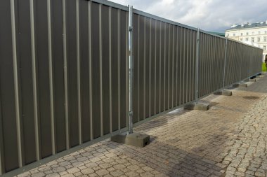 Modern gri bir çit şehir ortamında bir bariyer oluşturur, aşağıdaki kaldırıma gölgeler düşürür. İnşaat veya güvenlik temaları için ideal.