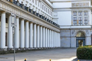 Beyaz sütunları ve süslü detayları olan klasik mimari bir binanın detaylı bir görüntüsü. Yürüyüş yolu boş, yapının zarafetini gösteriyor..
