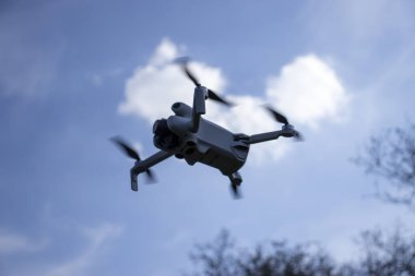 Yumuşak bulutlu mavi gökyüzüne karşı havada yakalanmış uçan bir dron. İnsansız hava aracı odak noktasında bulanık bir ağaç zeminiyle pervanelerini hareket ettiriyor..