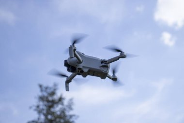 Uçan bir dron, pervanelerde hareket bulanıklığı olan açık mavi bir gökyüzüne karşı havada yakalandı. Arka planda, İHA 'nın açık havada kullanıldığını gösteren bulanık bir ağaç var..