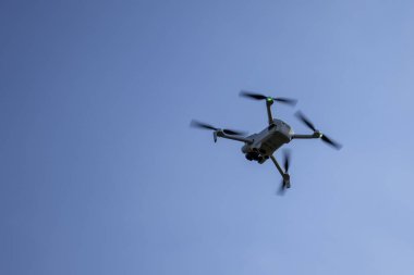 Beyaz dron açık mavi bir arka planda gökyüzünde uçuyor. İnsansız hava aracı pervanelerinde yeşil ışık yakıyor. Teknoloji ve hava fotoğrafçılığı temaları için ideal.