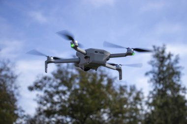 Arka planda bulanık ağaçlarla gökyüzünde uçan bir dron. İHA 'nın pervaneleri dönüyor ve yeşil navigasyon ışıkları yanıyor. Mavi gökyüzüne karşı insansız hava aracına odaklanıyoruz.