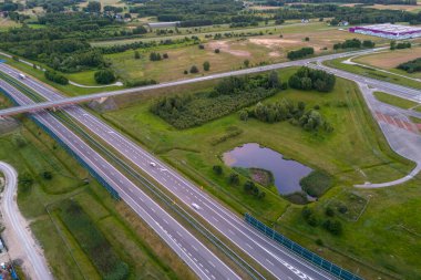 Yeşil alanlar ve ağaçlarla çevrili bir otoyol kavşağının havadan görüntüsü. Arabalar ses engellerinin yanı sıra yolda da görülebilir. Görüntü huzurlu bir kırsal yerleşim gösteriyor..