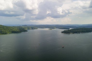 Bulutlu bir gökyüzünün altında yemyeşil tepelerle çevrili sakin bir göl manzarası. Tekneler, doğanın güzelliğini gösteren sularda görülebilir..