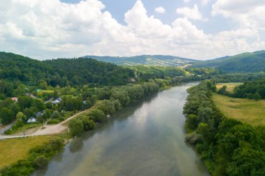 Parlak mavi gökyüzünün altında yemyeşil ve tepelerle çevrili sakin bir nehrin havadan görünüşü.
