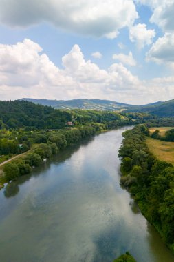 Tüylü bulutlarla dolu mavi gökyüzünün altında, yemyeşil tepelerde süzülen huzurlu bir nehrin havadan görünüşü.