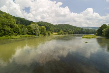 Tüylü beyaz bulutlarla dolu masmavi bir gökyüzünün altında yemyeşil ormanlardan akan bir nehrin huzurlu manzarası.
