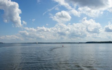 Sakin sularda süzülen yelkenlilerin ve parlak mavi gökyüzüne dağılmış kabarık bulutların olduğu sakin bir göl sahnesi. Doğa ve açık hava temaları için ideal.