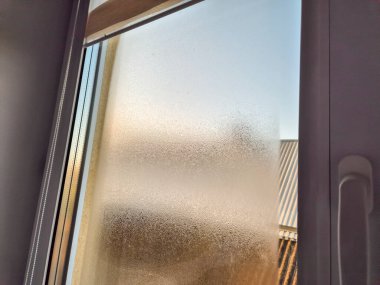 Dışarıdaki manzarayı aydınlatan yumuşak bir sabah ışığı olan buzlu cam bir pencere. Yoğunlaşma resme derinlik ve sıcaklık ekleyen soyut bir doku yaratır..