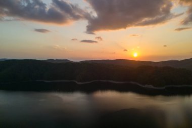 Arka planda tepeler olan sakin bir gölün üzerinde çarpıcı bir gün batımı. Alacakaranlıkta doğanın güzelliğini yakalamak için mükemmel bir an..