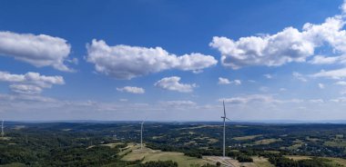 Tüylü bulutlarla dolu canlı mavi bir gökyüzünün altında yemyeşil bir arazide rüzgar türbinlerinin hava manzarası. Yenilenebilir enerji kavramı.