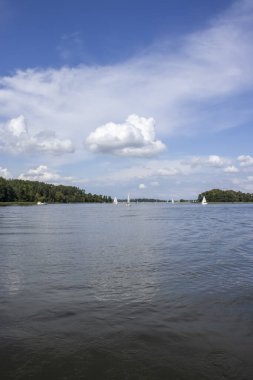 Parlak mavi bir gökyüzünün altında süzülen yelkenlilerin olduğu sakin bir göl manzarası. Rahatlamak ve doğa aşıkları için mükemmel..