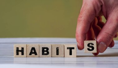 Kişisel gelişim ve değişim kavramını sembolize eden, açık ahşap bir yüzeye 'HABITS' kelimesinin önüne bir harf bloğu koyan bir el..