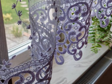 Zarif mor dantelli perde parlak bir pencereyi çerçeveliyor ve iç dekora zarif bir dokunuş ekliyor. Yumuşak bir sabah ışığı altında, kumaşın dokusunu ve detaylarını vurguluyor..