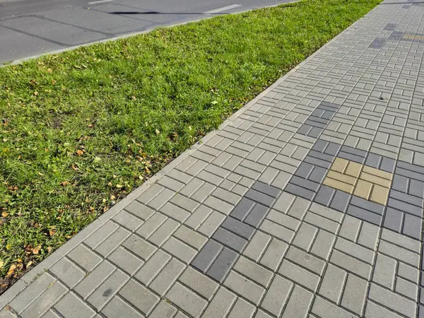 Şehir manzarasını vurgulayan yeşil çimlerin yanında desenli fayansların yer aldığı modern bir kaldırımın yakın çekim görüntüsü