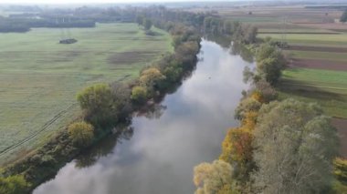 Açık arazilerle çevrili, her iki taraftaki ağaçları yansıtan sakin bir nehrin havadan görünüşü..