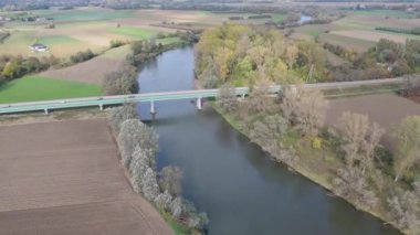 Kırsal alanda yeşil ve kahverengi tarlalı bir nehirden geçen bir köprünün insansız hava aracı görüntüsü.