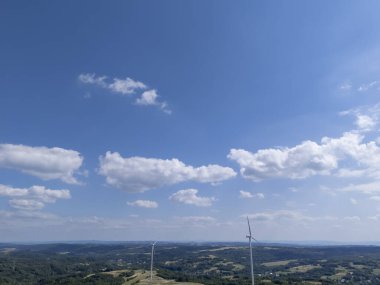 Pofuduk bulutlar ve ufuktaki rüzgar türbinleri tarafından vurgulanan parlak mavi gökyüzünün altındaki yuvarlanan tepelerin nefes kesici görüntüsü doğanın güzelliğini ve yenilenebilir enerjiyi yakalıyor..