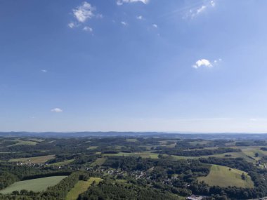 Parlak mavi gökyüzünün altındaki yemyeşil tarlaların ve yuvarlanan tepelerin panoramik görüntüsü. Manzara sakin ve doğanın güzelliğini gözler önüne seriyor.