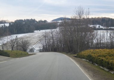 Sakin bir kış manzarası karlı tepeler ve bulutlu bir gökyüzünün altındaki ağaçlar arasında dolambaçlı bir yol gösteriyor..