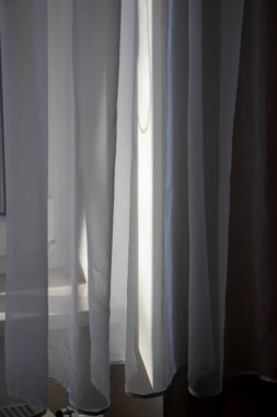 Saf beyaz perdelerin yakın çekimi meltemde hafifçe sallanarak yumuşak güneş ışığının sakin ve rahatlatıcı bir atmosfer yaratmasını sağlar.