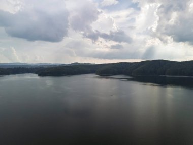 Bulutlu bir gökyüzünün altında yemyeşil ormanlarla çevrili sakin bir göl manzarası dinlenmek ve ilham vermek için huzurlu doğal bir manzara sunuyor..