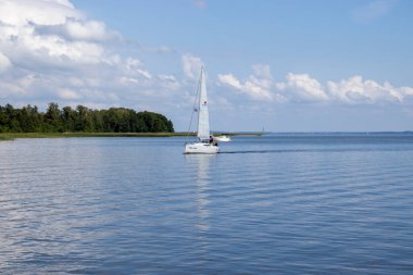 Mikolajki Polonya - 29 Eylül 2024: Bereketli yeşillikler ve mavi gökyüzüyle çevrili sakin sularda süzülen bir yelkenli manzarası.