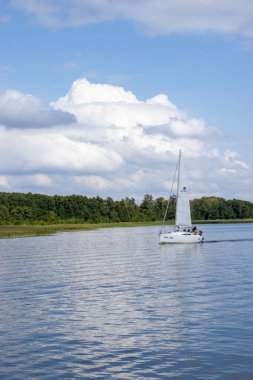 Mikolajki Polonya - 29 Eylül 2024: Bereketli yeşil ağaçlar ve parlak mavi gökyüzünde kabarık bulutlarla çevrili sakin sularda süzülen bir yelkenlinin sakin manzarası.