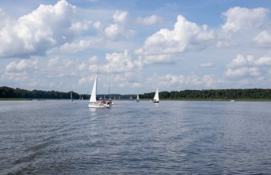 Mikolajki Polonya - 29 Eylül 2024: Mavi gökyüzü ve açık hava meraklıları ve doğa severler için mükemmel beyaz bulutlarla sakin bir göl üzerinde gezinen yelkenlilerin manzarası.