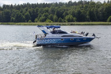 Mikolajki Polonya - 29 Eylül 2024: Bereketli yeşil ağaçlarla çevrili sakin sularda gezinen mavi bir tekne yaz tatili aktivitelerinin özünü yakalıyor.